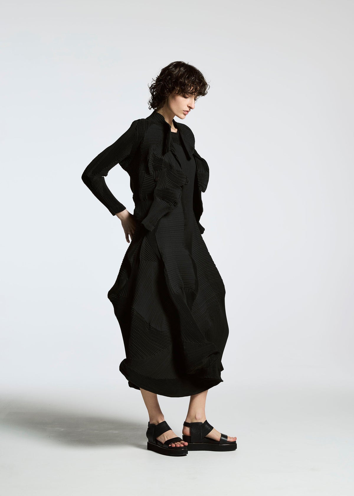 A person with short curly hair stands against a white background, wearing a long textured black dress and the TYPE-Ⅰ 008 jacket by A-POC ABLE ISSEY MIYAKE, paired with black platform sandals. They look down with hands on hips. view 3 of 4