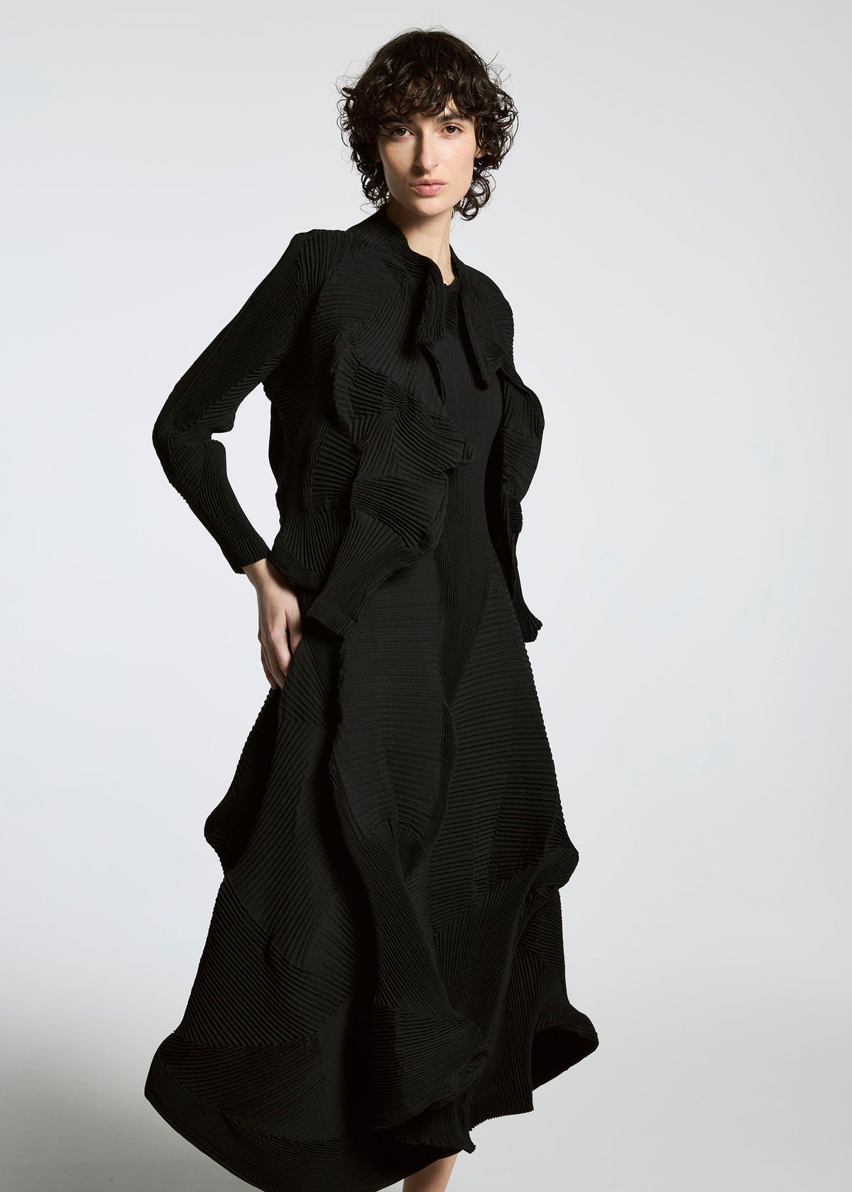 A person with short curly hair wears the TYPE-Ⅰ 008 dress by A-POC ABLE ISSEY MIYAKE and a sleek Steam Stretch jacket, standing against a plain light background. view 4 of 4