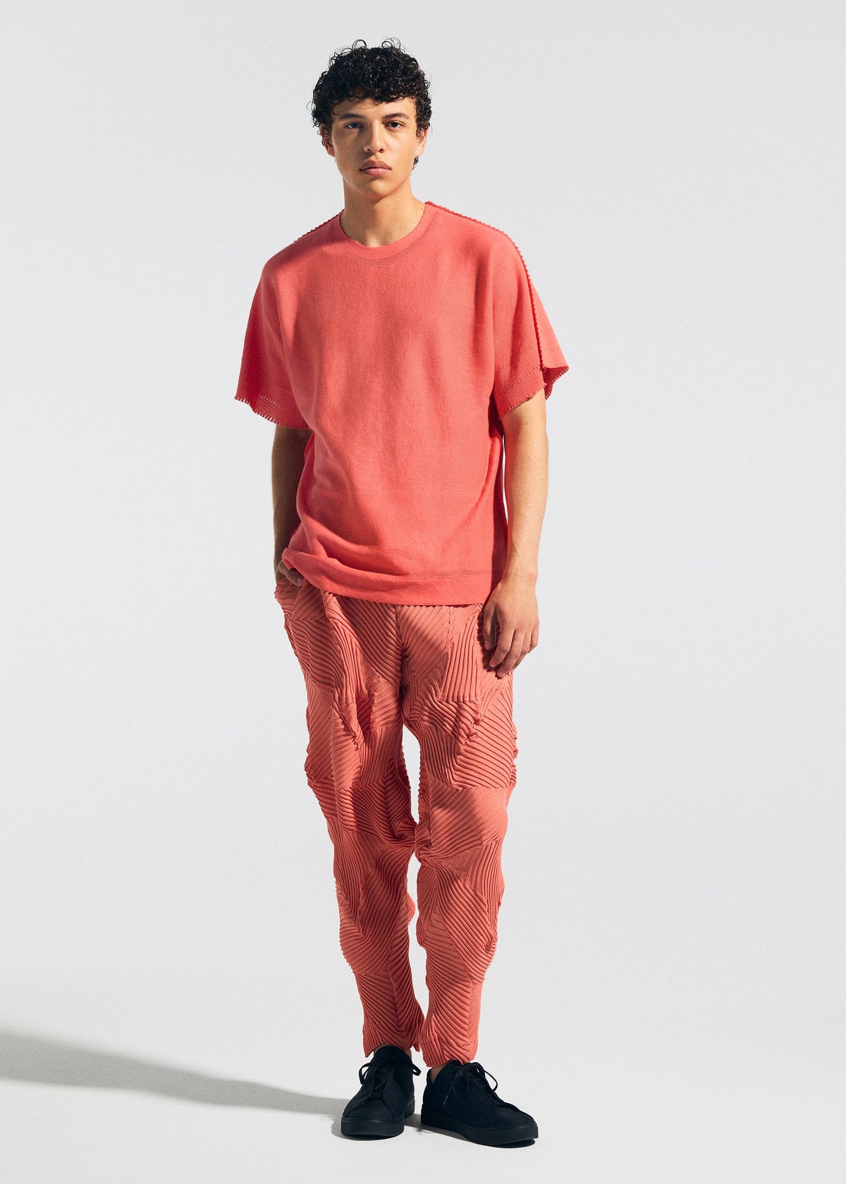 Wearing the TYPE-P 003-1 set by A-POC ABLE ISSEY MIYAKE, an individual with short, curly hair poses against a plain background. The loose-fit coral pink top and matching pleated pants offer versatile everyday comfort, styled with black shoes. view 6 of 9