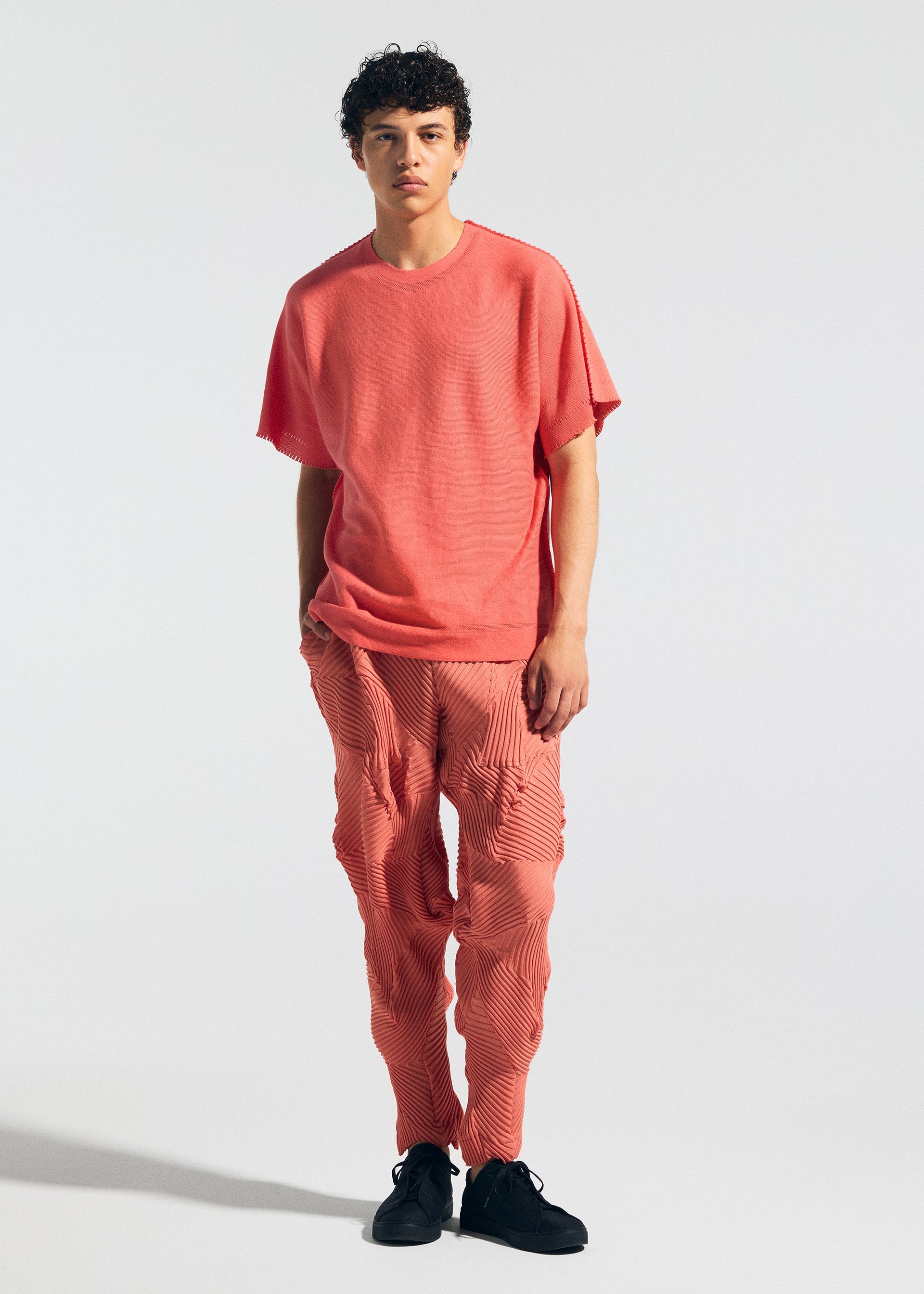 Zoomed view Wearing the TYPE-P 003-1 set by A-POC ABLE ISSEY MIYAKE, an individual with short, curly hair poses against a plain background. The loose-fit coral pink top and matching pleated pants offer versatile everyday comfort, styled with black shoes.