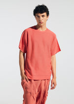 A young person with short curly hair wears the TYPE-P 003-1 set from A-POC ABLE ISSEY MIYAKE—loose, textured coral T-shirt and matching pants with Baked Stretch pleating—perfect for versatile everyday wear against a plain light background.