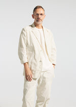A person with short hair and glasses wears the TYPE-U 001 off-white jacket, wrinkle-resistant pants, and a white shirt from A-POC ABLE ISSEY MIYAKE, standing against a plain light background and looking at the camera with a neutral expression.