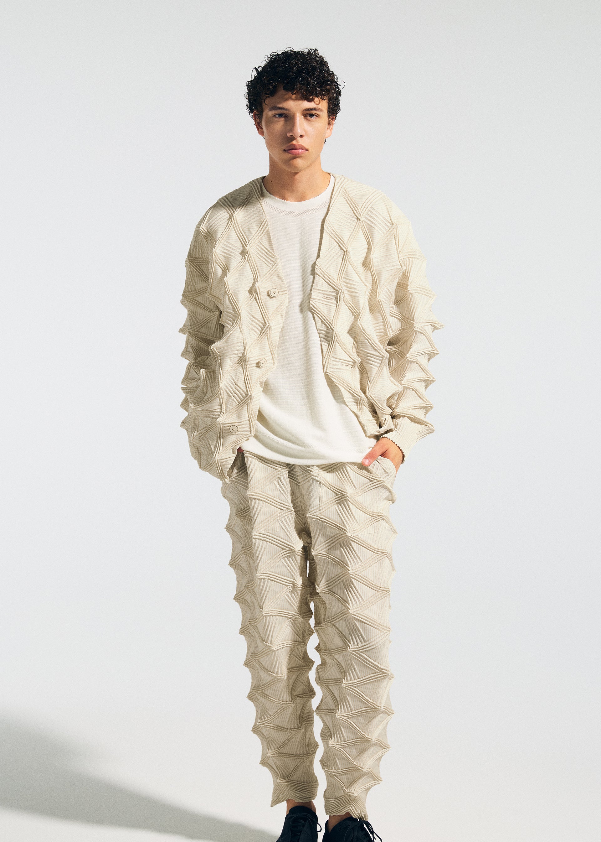 Zoomed view A person stands against a plain light background wearing the TYPE-O 023 set by A-POC ABLE ISSEY MIYAKE—a cream-colored ensemble featuring a geometric-textured jacket with three-dimensional sleeves and matching pants, styled with a white shirt and black shoes.