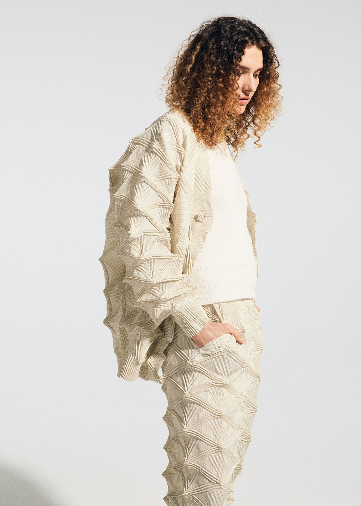 A person with curly hair wears a light-colored, textured outfit featuring geometric, 3D patterns on the jacket and pants, including the TYPE-O 023 from A-POC ABLE ISSEY MIYAKE—a Steam Stretch cardigan with three-dimensional sleeves—while standing against a plain background. view 7 of 10