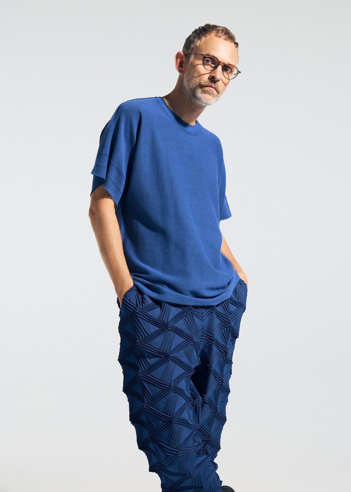 A man with short hair, glasses, and a beard wears the TYPE-O 023 knit t-shirt from A-POC ABLE ISSEY MIYAKE under a Steam Stretch cardigan with three-dimensional sleeves, paired with textured blue geometric-patterned pants. He stands with his hands in his pockets against a plain light background. view 8 of 10