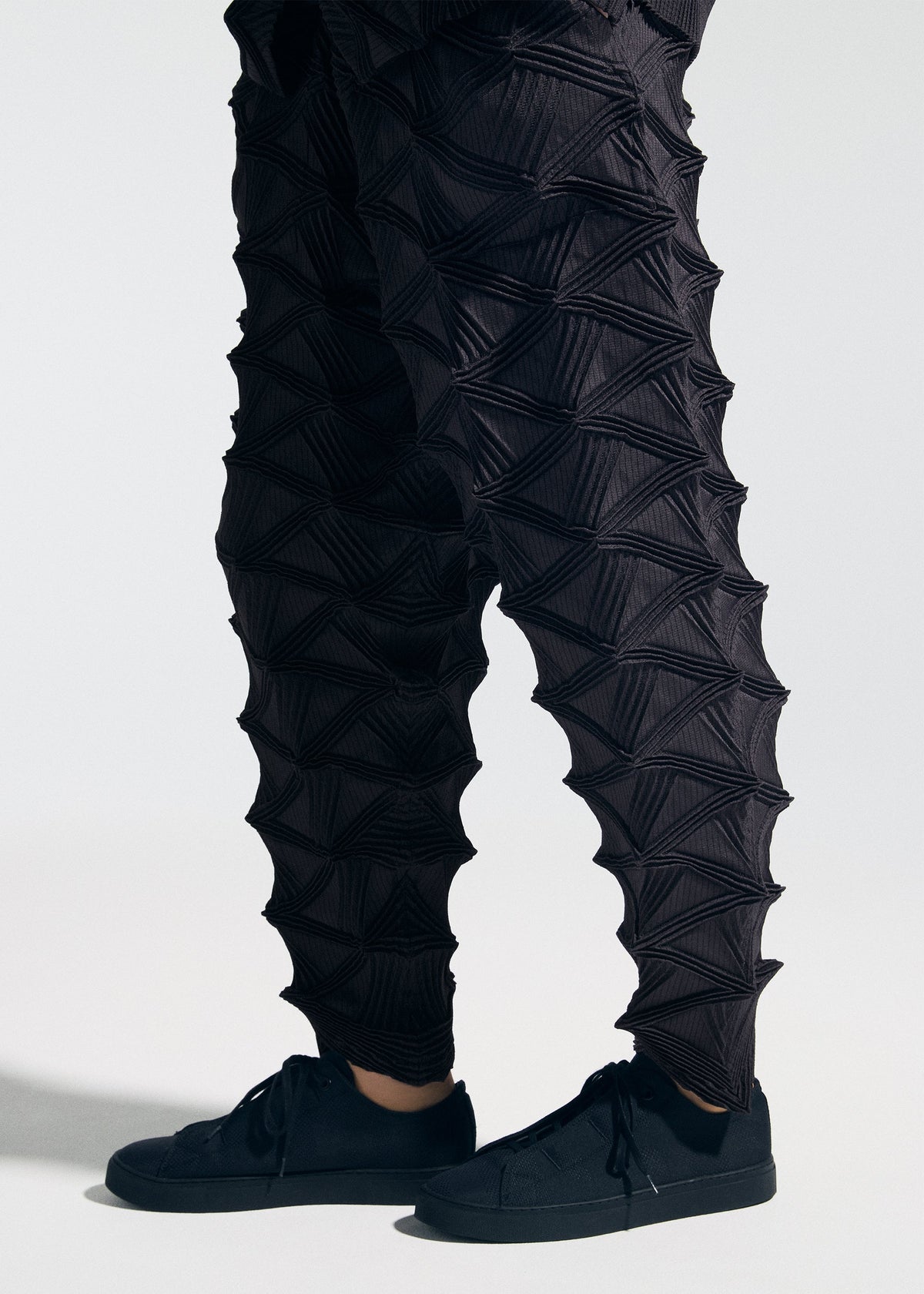 Wearing the A-POC ABLE ISSEY MIYAKE TYPE-O 023 sneakers and distinctive black pants featuring geometric, three-dimensional triangular textures, paired with a sleek pleated cardigan, this person stands against a plain light background. view 10 of 10