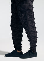 Wearing the A-POC ABLE ISSEY MIYAKE TYPE-O 023 sneakers and distinctive black pants featuring geometric, three-dimensional triangular textures, paired with a sleek pleated cardigan, this person stands against a plain light background.