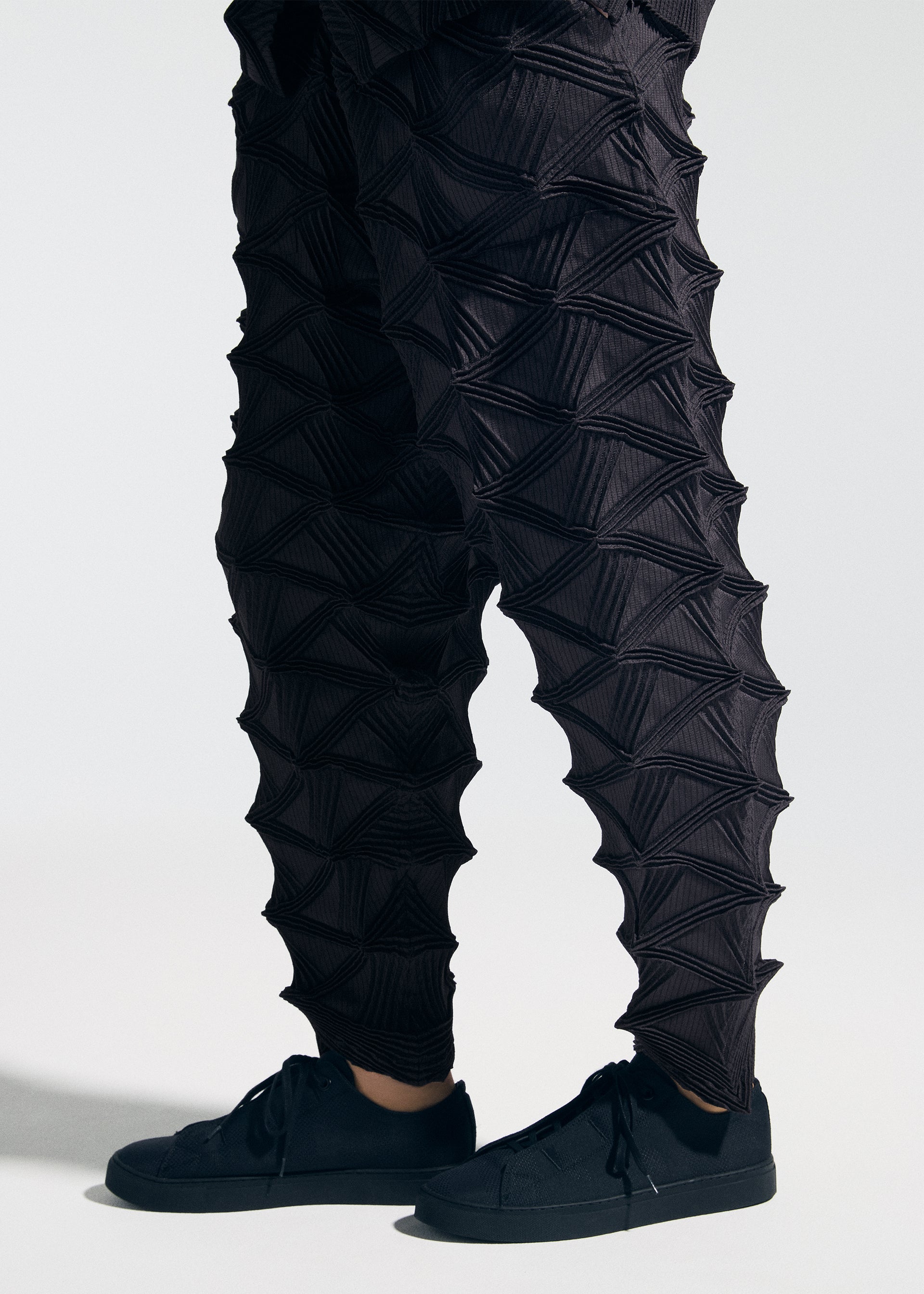 Zoomed view Wearing the A-POC ABLE ISSEY MIYAKE TYPE-O 023 sneakers and distinctive black pants featuring geometric, three-dimensional triangular textures, paired with a sleek pleated cardigan, this person stands against a plain light background.