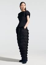 A person stands with hands in pockets, wearing the black TYPE-O 019 outfit by A-POC ABLE ISSEY MIYAKE, featuring a geometric 3D pattern made with the Steam Stretch technique on both top and zigzag pleat pants against a plain light background.