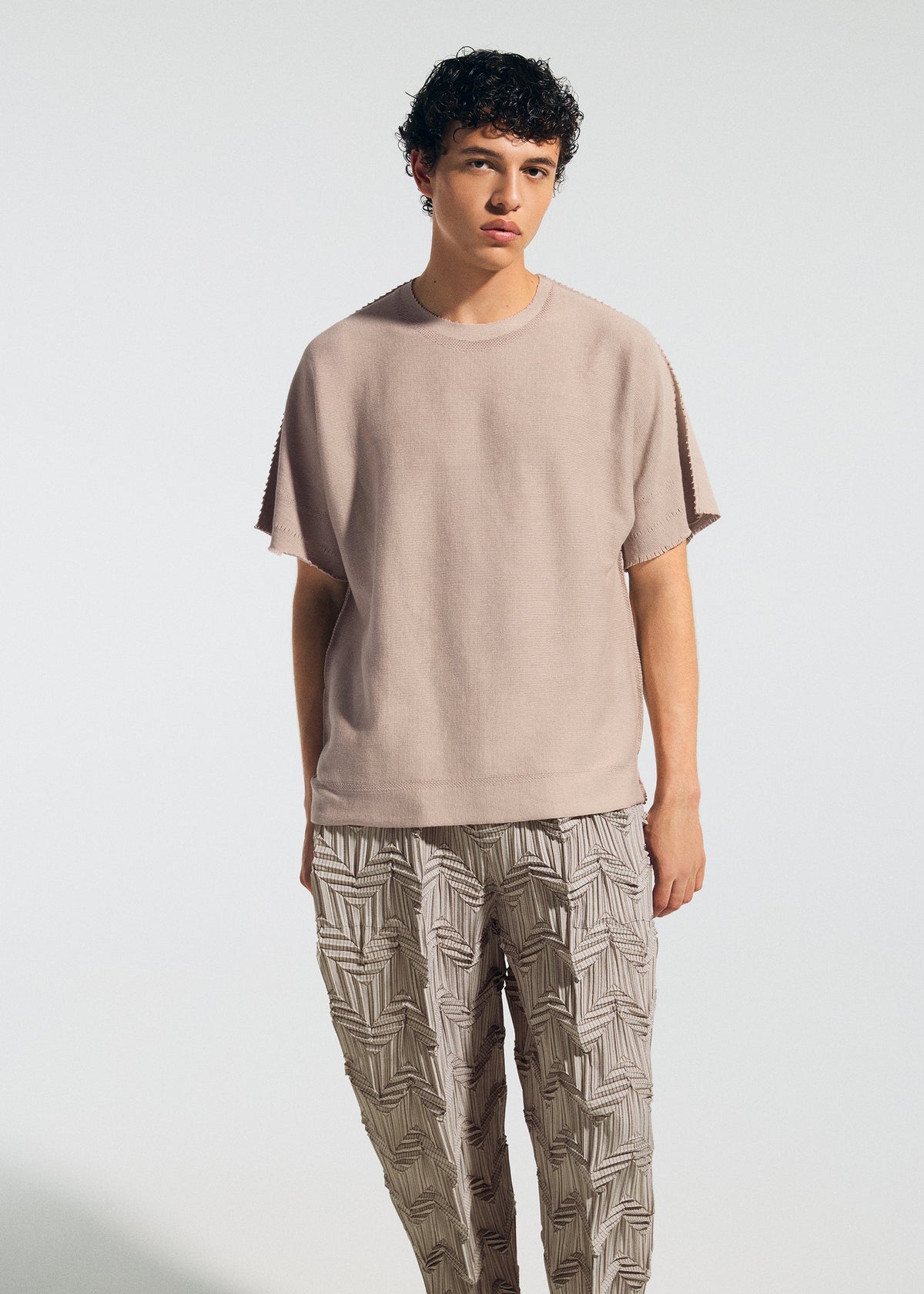 A young person with short curly hair wears the TYPE-O 019 zigzag pleat pants by A-POC ABLE ISSEY MIYAKE, featuring a three-dimensional pattern, paired with a loose beige top against a plain light background. view 8 of 11