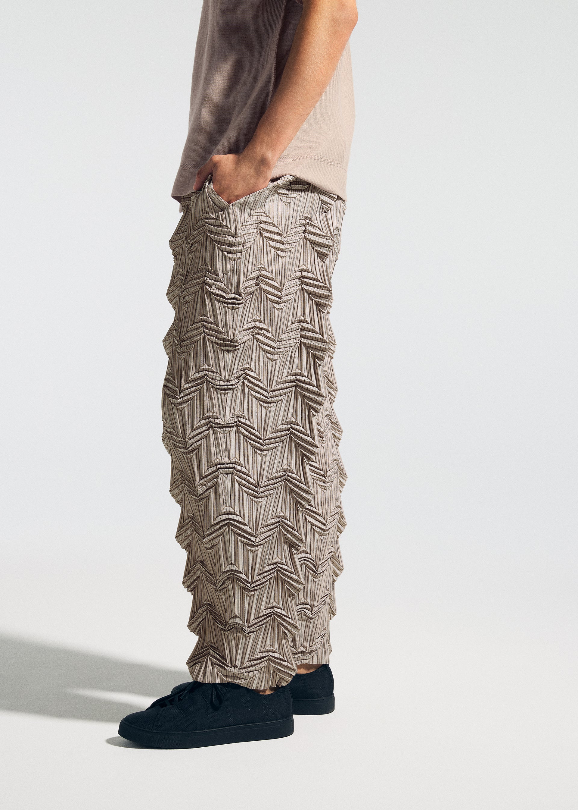 Zoomed view A person wears A-POC ABLE ISSEY MIYAKE TYPE-O 019 zigzag pleat pants with a 3D geometric design made by Steam Stretch, paired with a beige shirt and black sneakers, standing against a plain light background with hands in pockets.