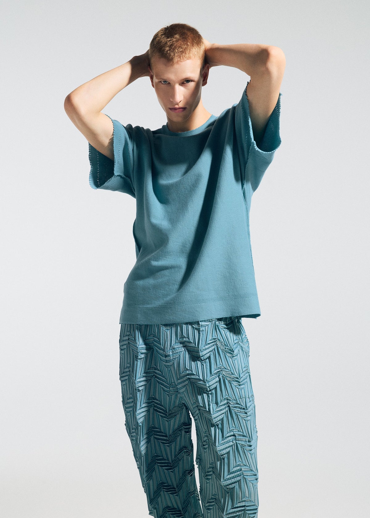 Wearing the TYPE-O 019 from A-POC ABLE ISSEY MIYAKE, a person poses with hands behind their head in a teal short-sleeve shirt and zigzag pleat pants, looking at the camera against a plain light background. view 10 of 11