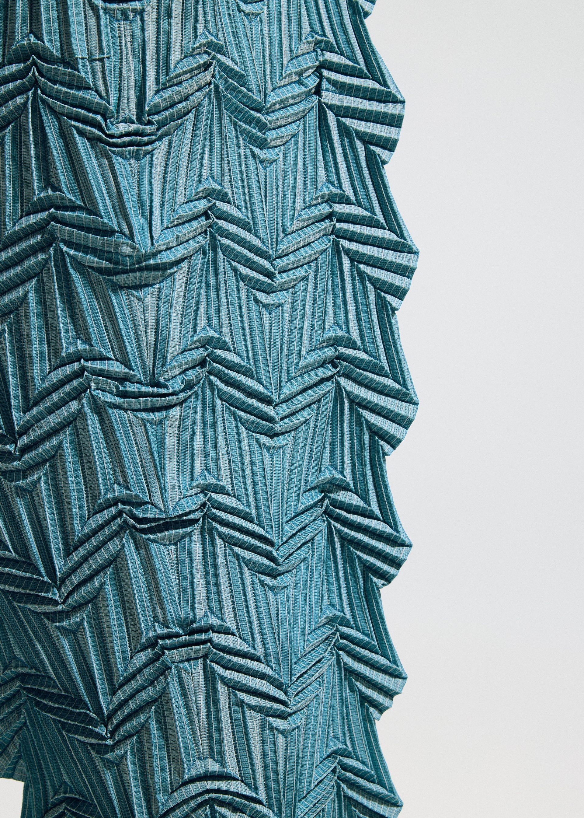 Zoomed view Close-up of TYPE-O 019 by A-POC ABLE ISSEY MIYAKE: blue fabric with geometric, zigzag, and layered patterns. The structured, three-dimensional effect resembles zigzag pleat pants crafted with the Steam Stretch technique.