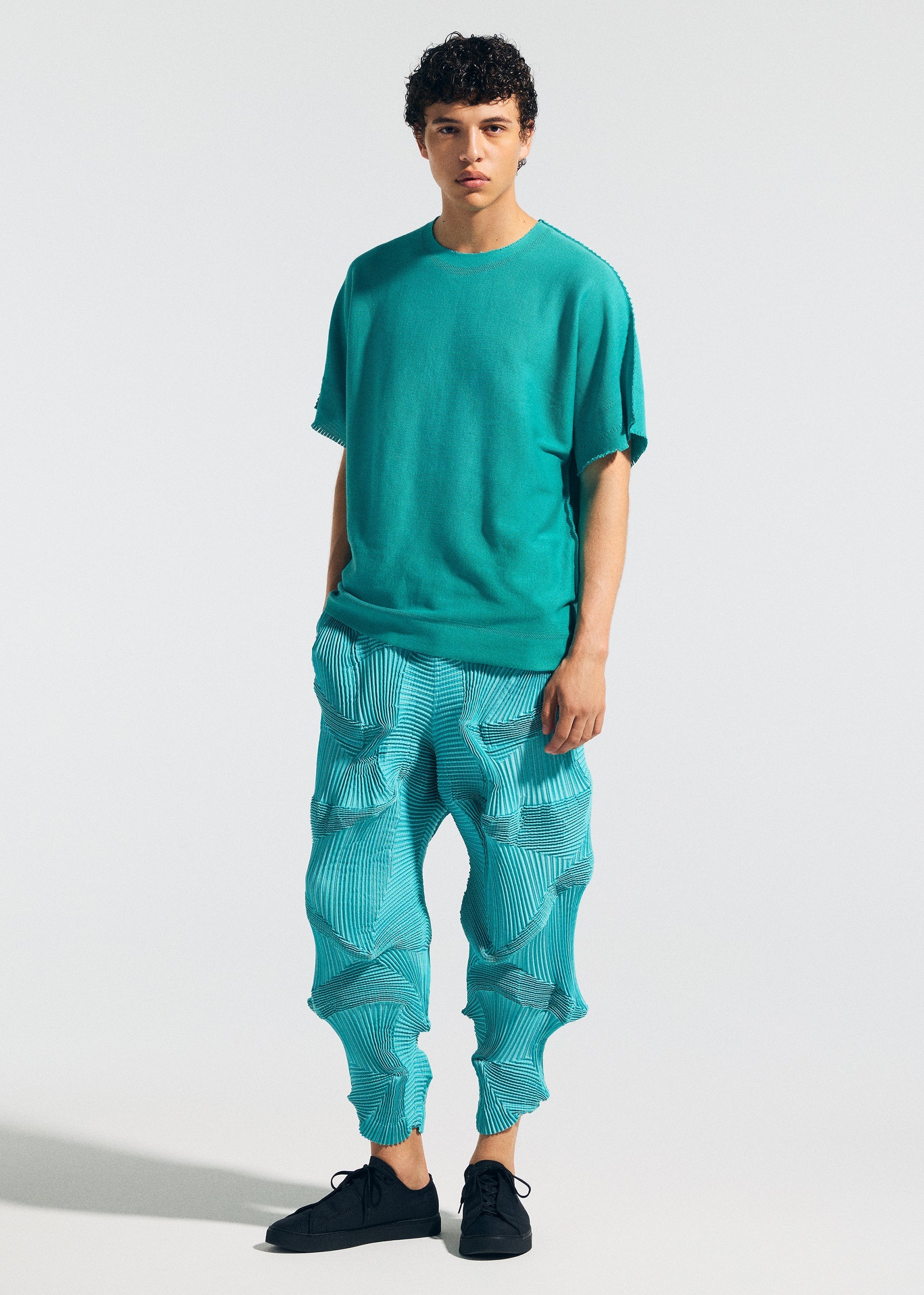 Zoomed view A young person faces forward against a plain light background, wearing A-POC ABLE ISSEY MIYAKE's TYPE-O 003: loose turquoise short-sleeve top and distinctive Steam Stretch pants with wavy sculpted patterns, paired with black shoes.