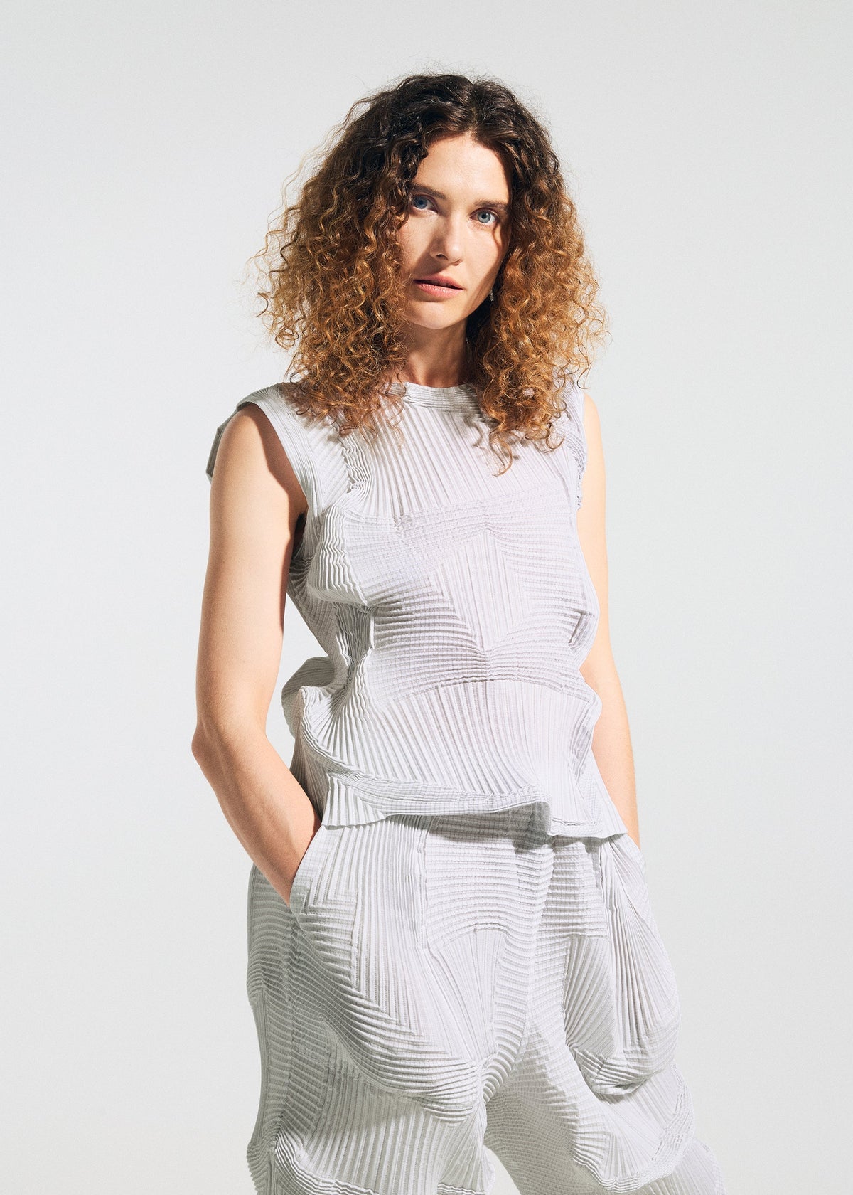 A woman with curly brown hair stands against a light background, wearing A-POC ABLE ISSEY MIYAKE's TYPE-O 003 sleeveless textured white top and matching Steam Stretch pants. Her hands are in her pockets as she faces the camera neutrally. view 8 of 8