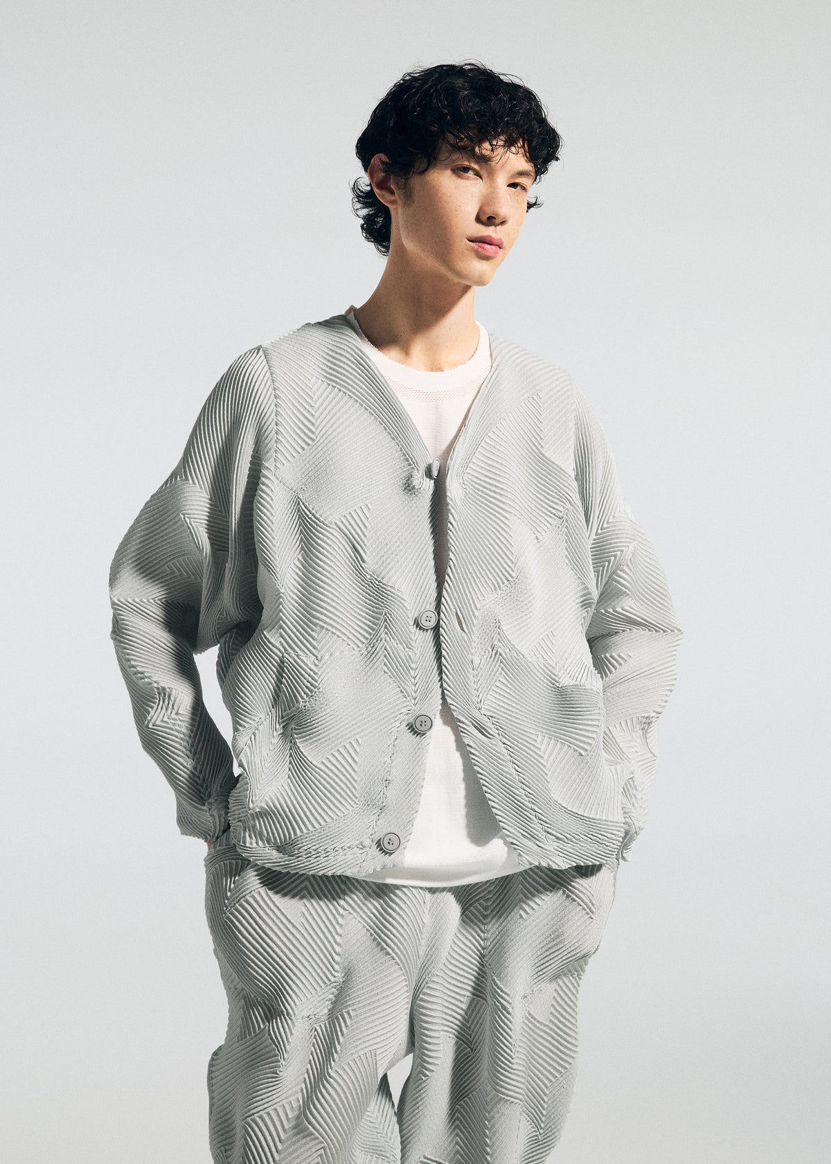 A person with short curly hair wears the TYPE-O 010 jacket and pleated pants by A-POC ABLE ISSEY MIYAKE—light gray, houndstooth patterned, over a white T-shirt—standing hands in pockets against a plain light background. view 6 of 8