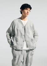 A person with short curly hair wears the TYPE-O 010 jacket and pleated pants by A-POC ABLE ISSEY MIYAKE—light gray, houndstooth patterned, over a white T-shirt—standing hands in pockets against a plain light background.