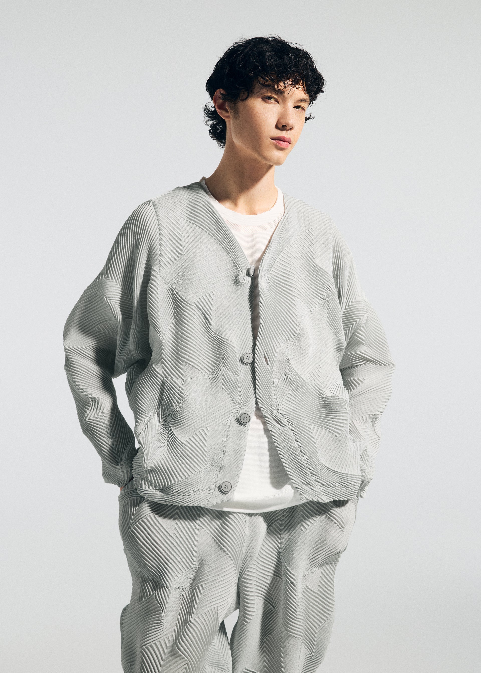 Zoomed view A person with short curly hair wears the TYPE-O 010 jacket and pleated pants by A-POC ABLE ISSEY MIYAKE—light gray, houndstooth patterned, over a white T-shirt—standing hands in pockets against a plain light background.