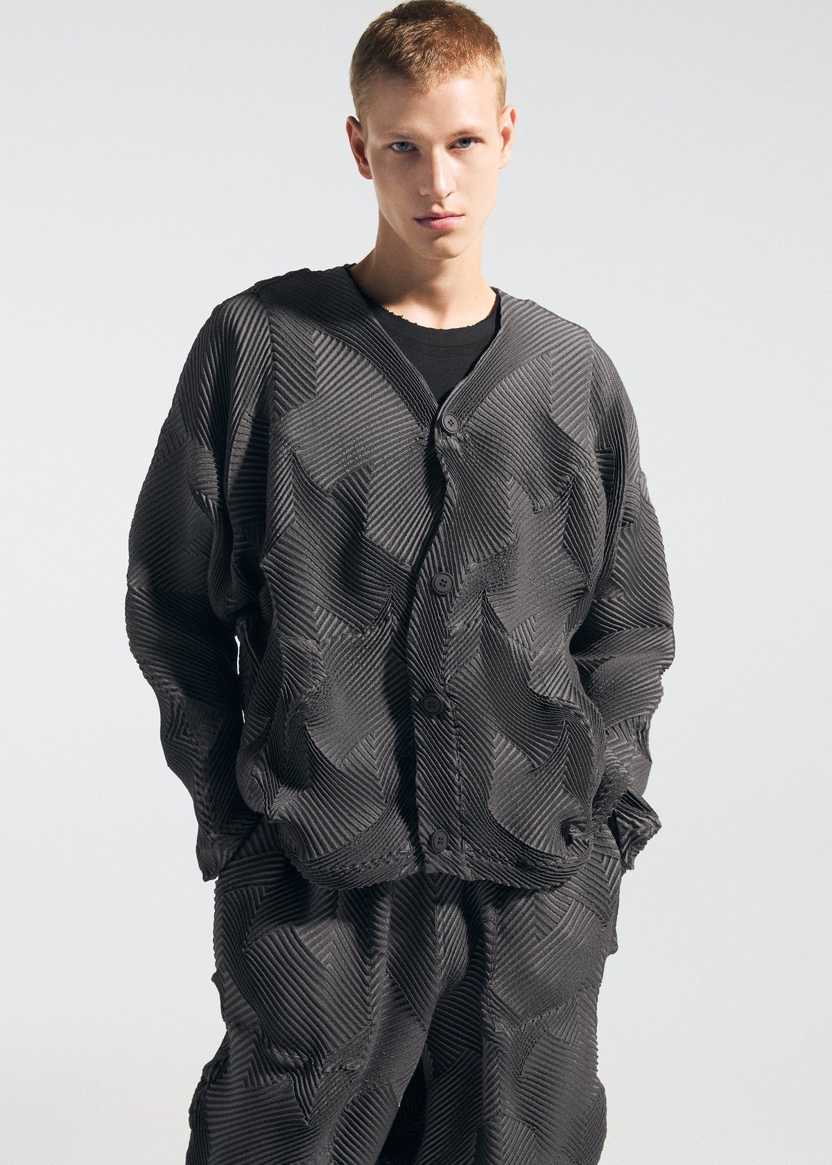 A person with short, light hair stands against a plain background, hands in pockets, wearing the TYPE-O 010 jacket and pleated houndstooth pants by A-POC ABLE ISSEY MIYAKE. view 8 of 8