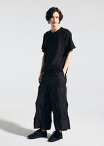 A person with short, wavy dark hair wears the A-POC ABLE ISSEY MIYAKE TYPE-W 009-1 loose black T-shirt and relaxed fit Steam Stretch pants, paired with black sneakers, standing against a plain white background with hands in pockets.
