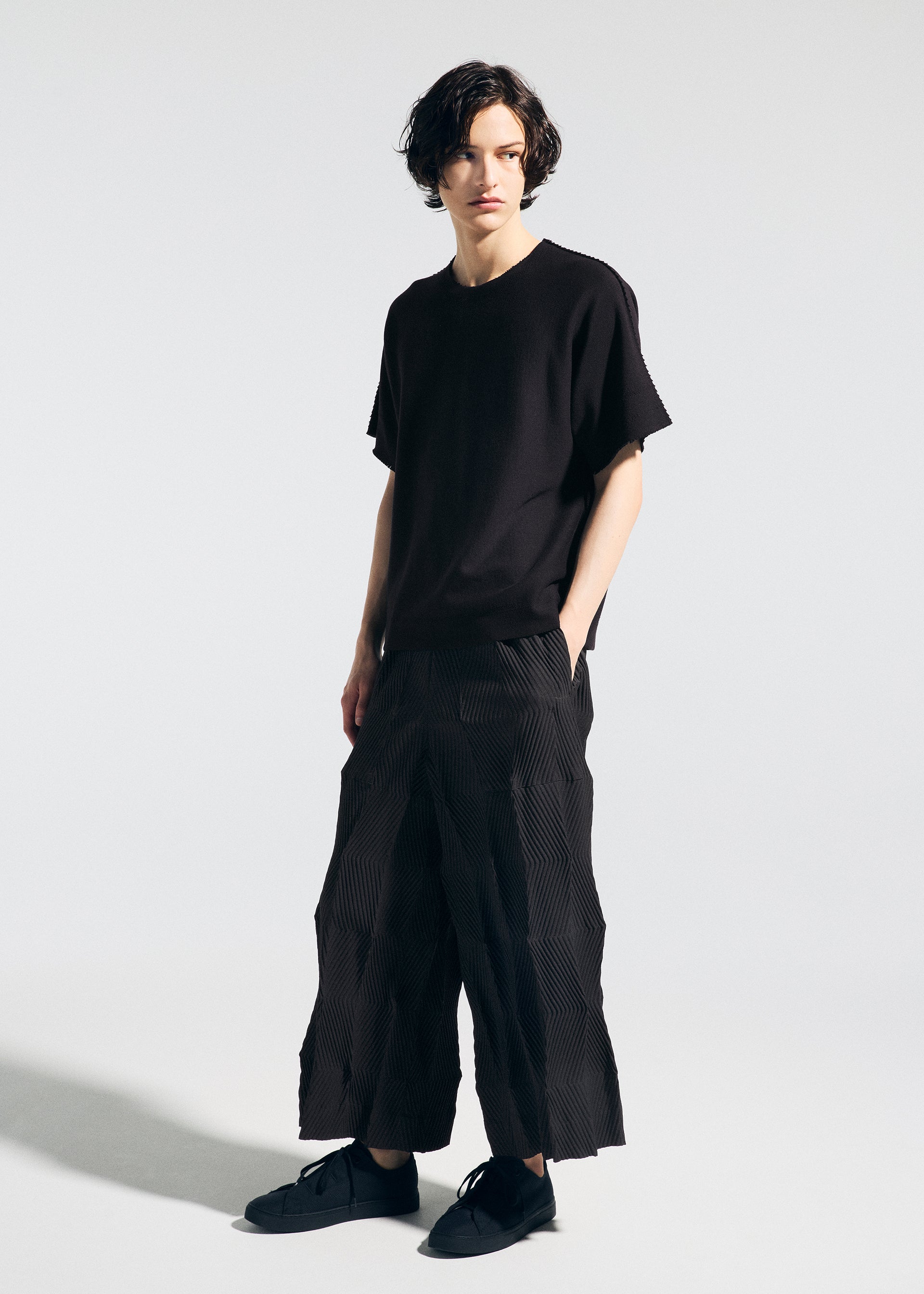 Zoomed view A person with short, wavy dark hair wears the A-POC ABLE ISSEY MIYAKE TYPE-W 009-1 loose black T-shirt and relaxed fit Steam Stretch pants, paired with black sneakers, standing against a plain white background with hands in pockets.