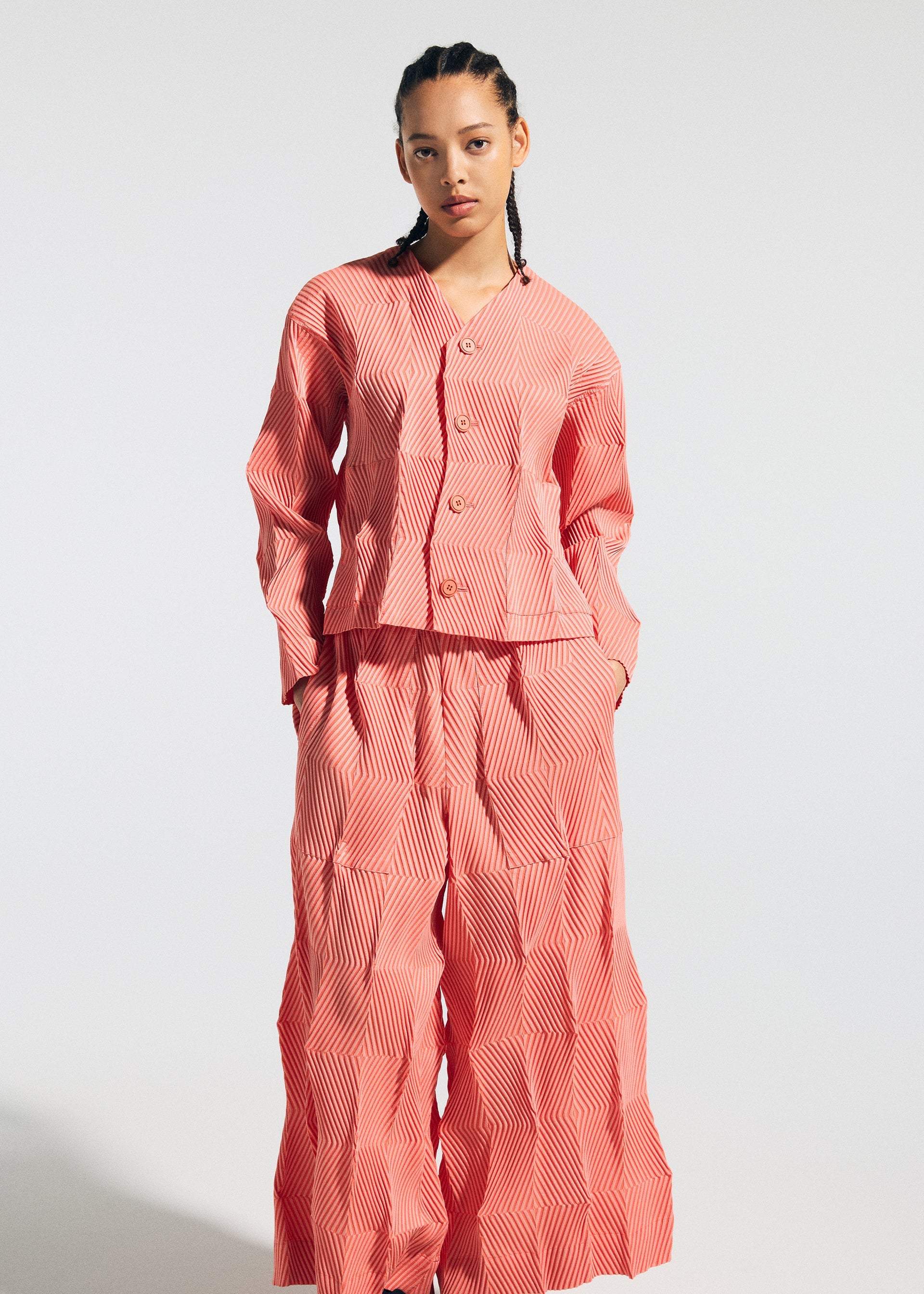 Zoomed view A person with braided hair wears the TYPE-W 009-1 button-up top and Steam Stretch pants by A-POC ABLE ISSEY MIYAKE, featuring a loose fit and geometric coral pink pattern, standing against a plain white background.