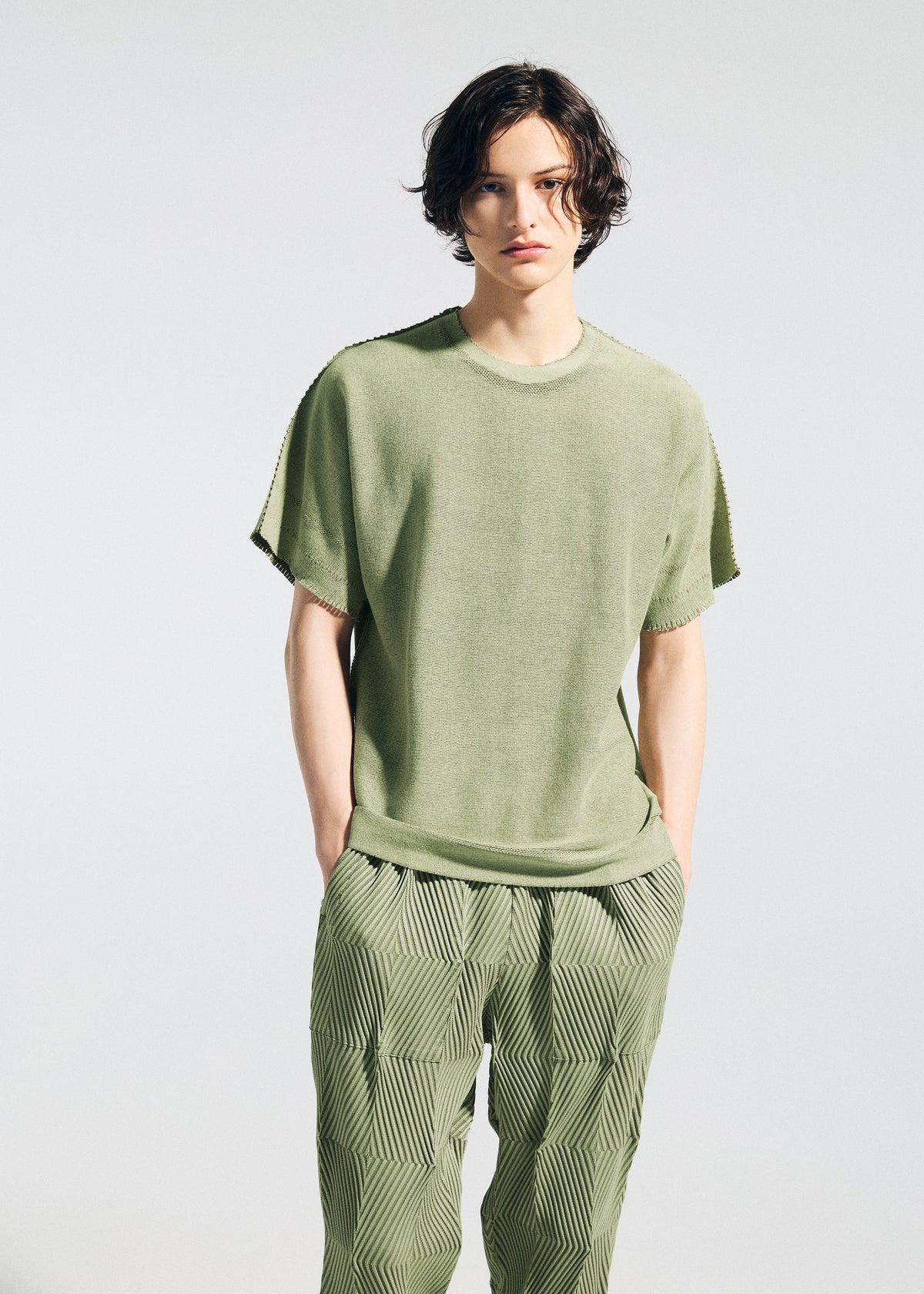 A person with wavy dark hair wears the TYPE-W 009-2 loose-fitting light green T-shirt and relaxed fit, patterned green Steam Stretch pants from A-POC ABLE ISSEY MIYAKE, standing against a plain light background. view 8 of 9