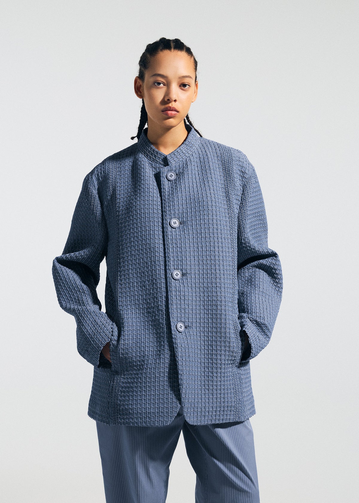 A person with braided hair models the TYPE-S 003 by A-POC ABLE ISSEY MIYAKE: a textured blue button-up jacket and pleated, lightweight stretchy pants, standing against a plain light background with hands in the jacket pockets. view 5 of 6