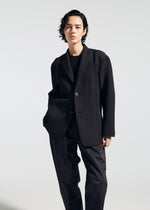 A person with short black hair models the A-POC ABLE ISSEY MIYAKE TYPE-S 003: a textured black blazer, black shirt, and loose accordion-pleat Steam Stretch pants, standing against a plain background with one hand in their pocket.