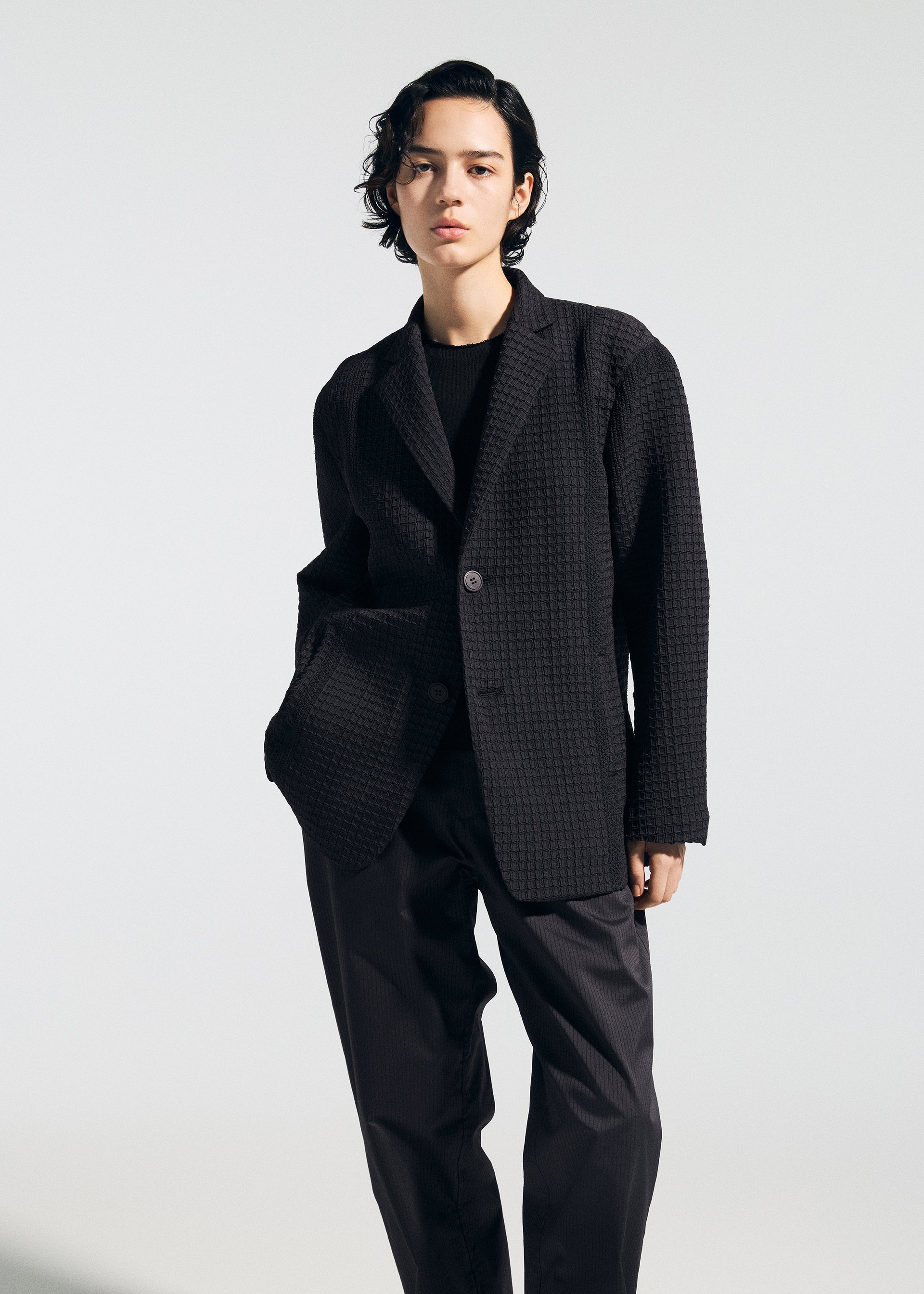 Zoomed view A person with short black hair models the A-POC ABLE ISSEY MIYAKE TYPE-S 003: a textured black blazer, black shirt, and loose accordion-pleat Steam Stretch pants, standing against a plain background with one hand in their pocket.