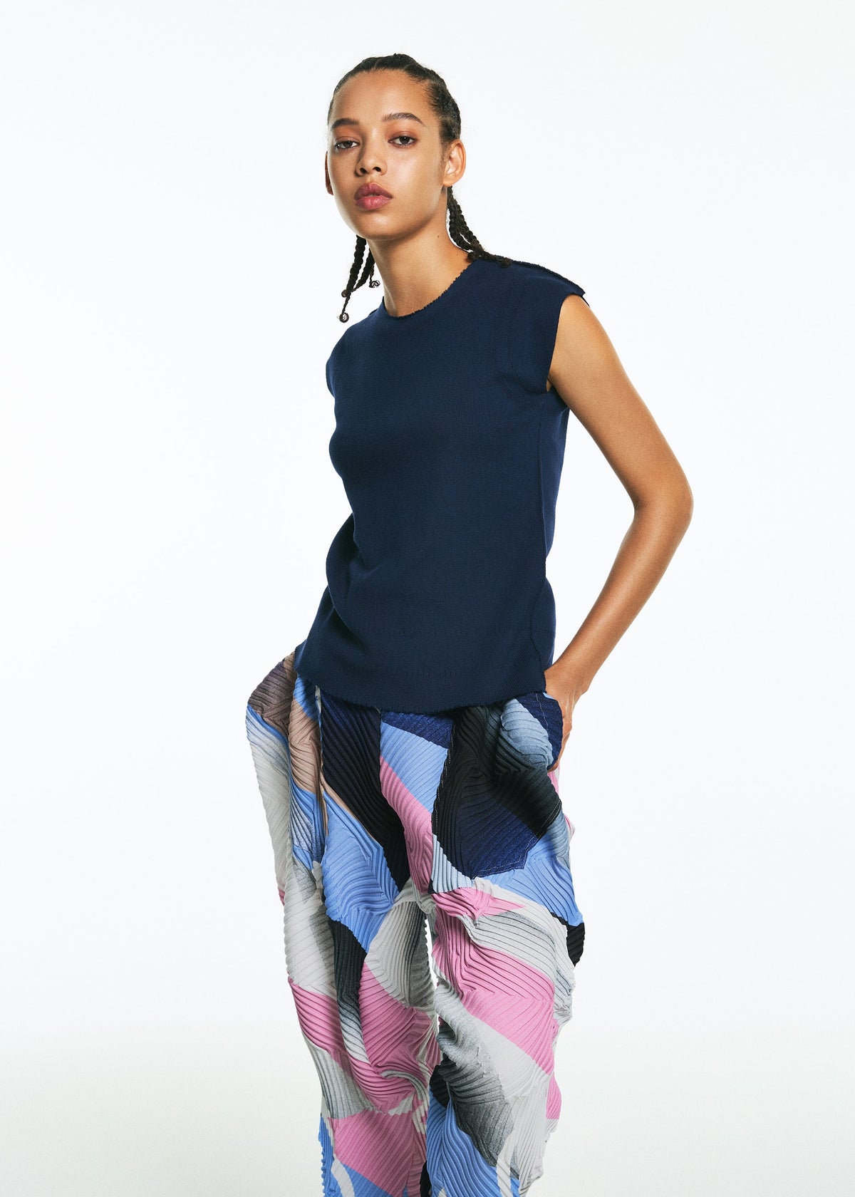 A woman with braided hair stands against a white background wearing the TYPE-XV SOAR NY project 001 pants by A-POC ABLE ISSEY MIYAKE, featuring loose pleats and a wavy pattern, her hands in her pockets. view 6 of 9