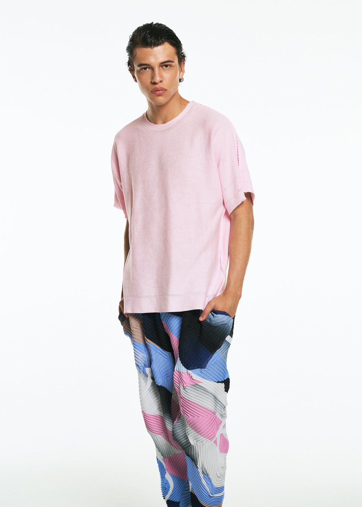 A young man with slicked-back hair models the TYPE-XV SOAR NY project 001 from A-POC ABLE ISSEY MIYAKE—wearing a loose light pink T-shirt and vibrant patterned pants in blue, pink, white, and black—against a plain white backdrop. view 7 of 9