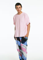 A young man with slicked-back hair models the TYPE-XV SOAR NY project 001 from A-POC ABLE ISSEY MIYAKE—wearing a loose light pink T-shirt and vibrant patterned pants in blue, pink, white, and black—against a plain white backdrop.