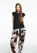A woman with long straight red hair wears the TYPE-XV SOAR NY project 001 from A-POC ABLE ISSEY MIYAKE: a black short-sleeve top and patterned wide-leg pants, standing against a plain white background.