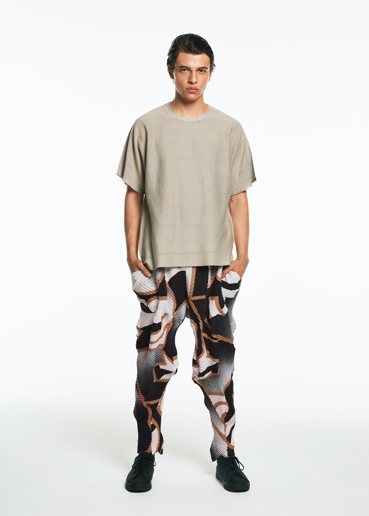 A person stands against a white background wearing the TYPE-XV SOAR NY project 001 by A-POC ABLE ISSEY MIYAKE: a loose beige short-sleeve top, abstract black, white, and brown patterned pants, and black shoes, with hands in their pockets. view 9 of 9