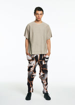 A person stands against a white background wearing the TYPE-XV SOAR NY project 001 by A-POC ABLE ISSEY MIYAKE: a loose beige short-sleeve top, abstract black, white, and brown patterned pants, and black shoes, with hands in their pockets.