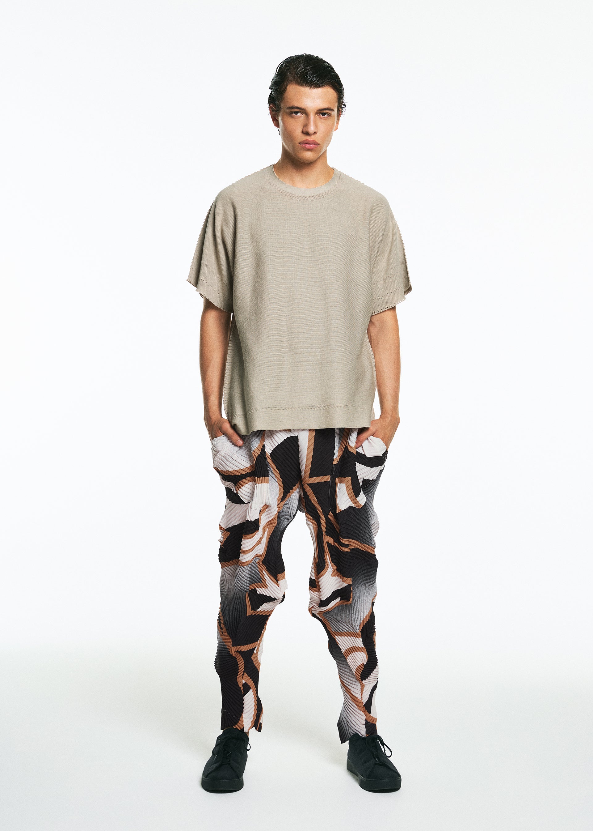Zoomed view A person stands against a white background wearing the TYPE-XV SOAR NY project 001 by A-POC ABLE ISSEY MIYAKE: a loose beige short-sleeve top, abstract black, white, and brown patterned pants, and black shoes, with hands in their pockets.