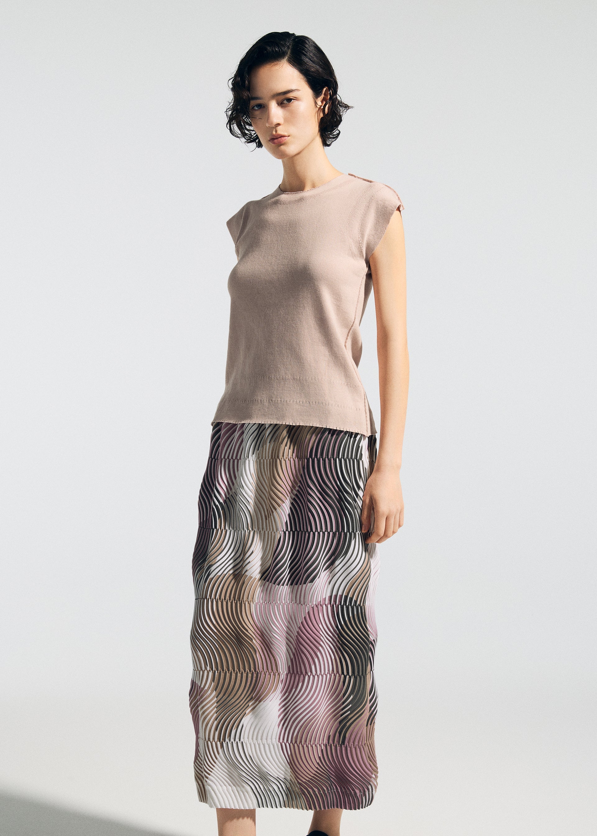 Zoomed view A woman with short dark hair wears a sleeveless beige top and the TYPE-P 005 Baked Stretch skirt by A-POC ABLE ISSEY MIYAKE, featuring a wavy texture in neutral tones, standing against a plain light background.