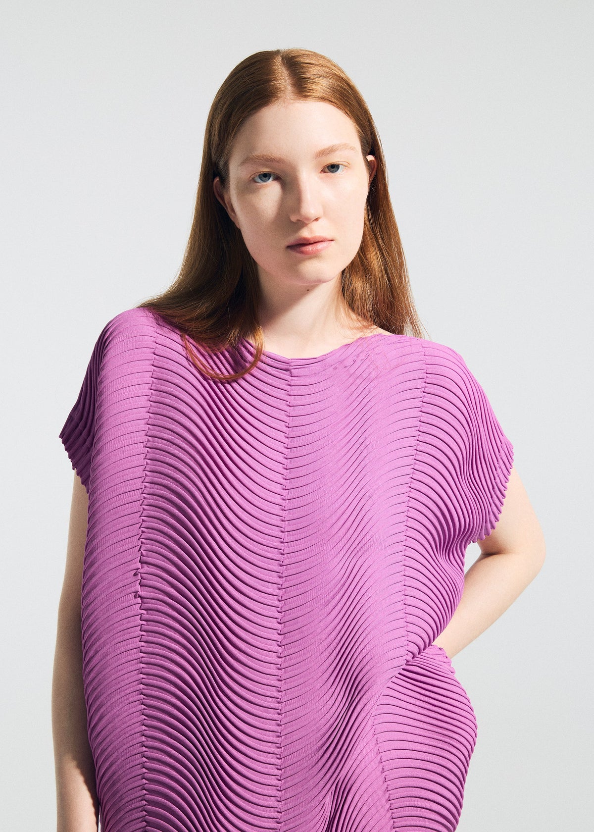 A woman with long, straight auburn hair wears the TYPE-P 006 Baked Stretch top by A-POC ABLE ISSEY MIYAKE—a bright purple piece with horizontal pleats. She stands against a plain light background, gazing forward with a neutral expression. view 7 of 8