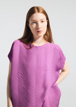 A woman with long, straight auburn hair wears the TYPE-P 006 Baked Stretch top by A-POC ABLE ISSEY MIYAKE—a bright purple piece with horizontal pleats. She stands against a plain light background, gazing forward with a neutral expression.