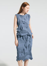 A woman with long red hair stands against a plain background, wearing the TYPE-P 003-2 sleeveless blue top and versatile pleated skirt by A-POC ABLE ISSEY MIYAKE. She looks slightly down and to the side.