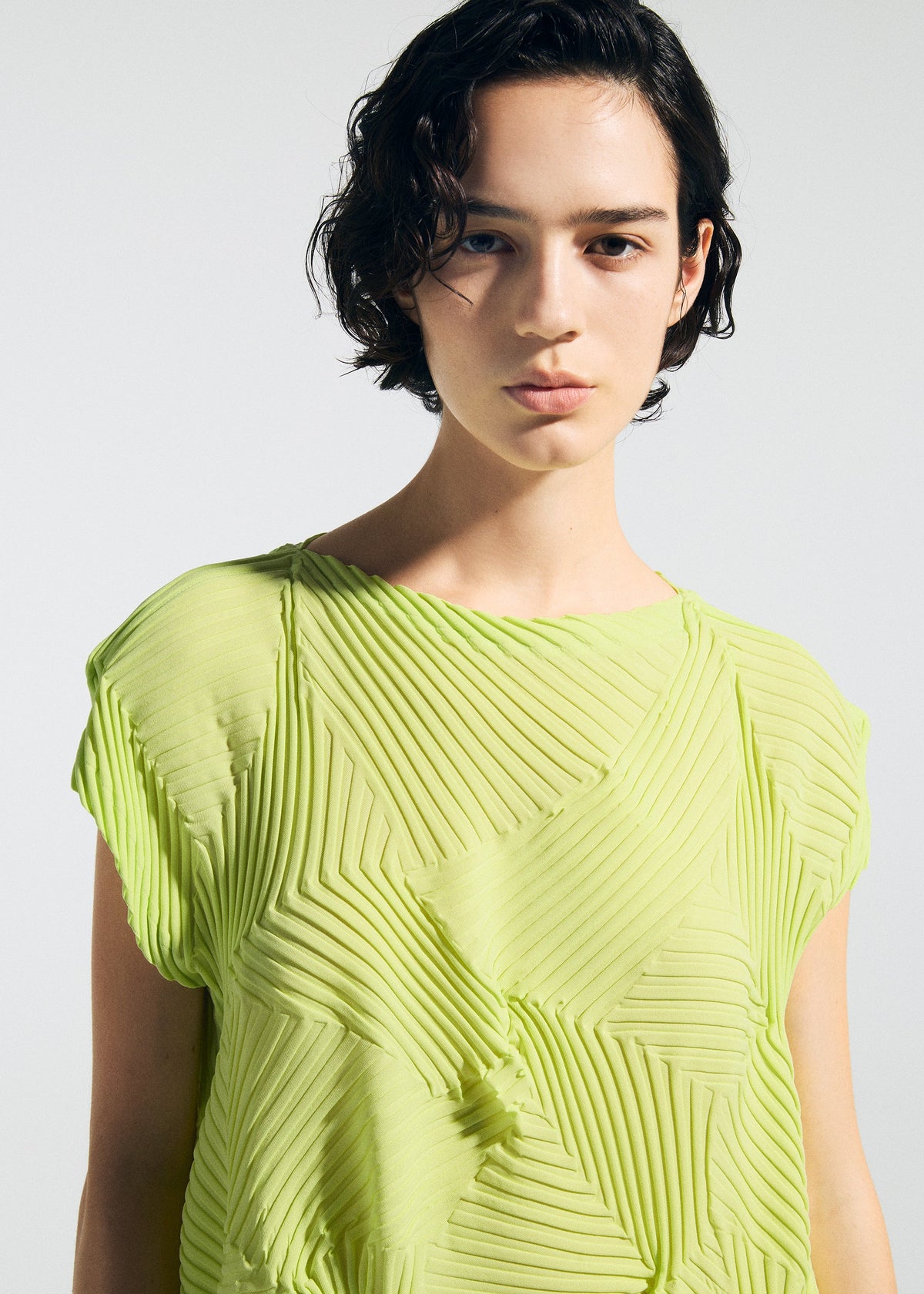A person with short, dark, wavy hair models the TYPE-P 003-2 top by A-POC ABLE ISSEY MIYAKE—a light green textured piece with geometric patterns—ideal for versatile everyday wear. A plain background highlights their direct gaze and neutral expression. view 6 of 8