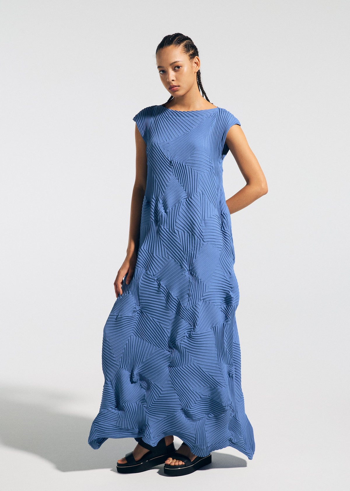 A model in the TYPE-P 003-1 dress by A-POC ABLE ISSEY MIYAKE stands against a plain light background, wearing a blue, floor-length pleated dress with geometric patterns, cap sleeves, and a relaxed fit. view 5 of 7