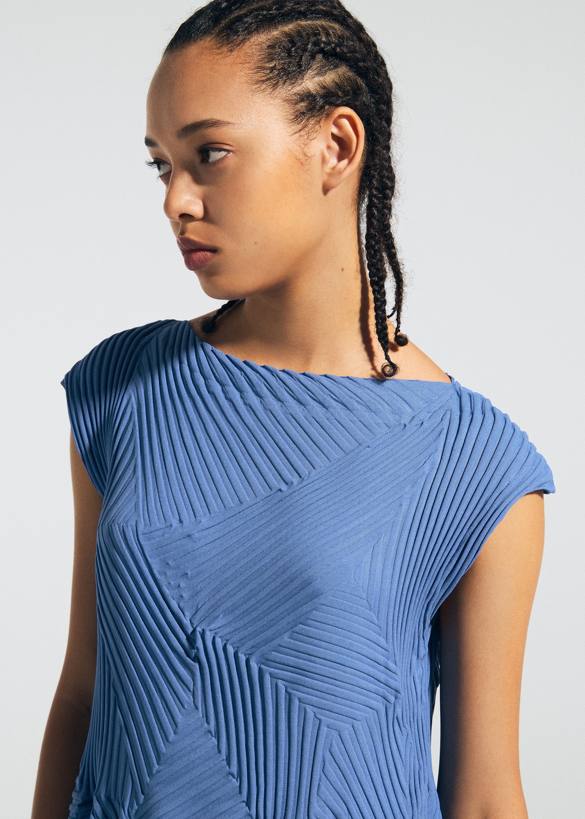 Zoomed view A person with braided hair wears the TYPE-P 003-1 textured blue pleated dress by A-POC ABLE ISSEY MIYAKE, looking to the side against a plain light background.