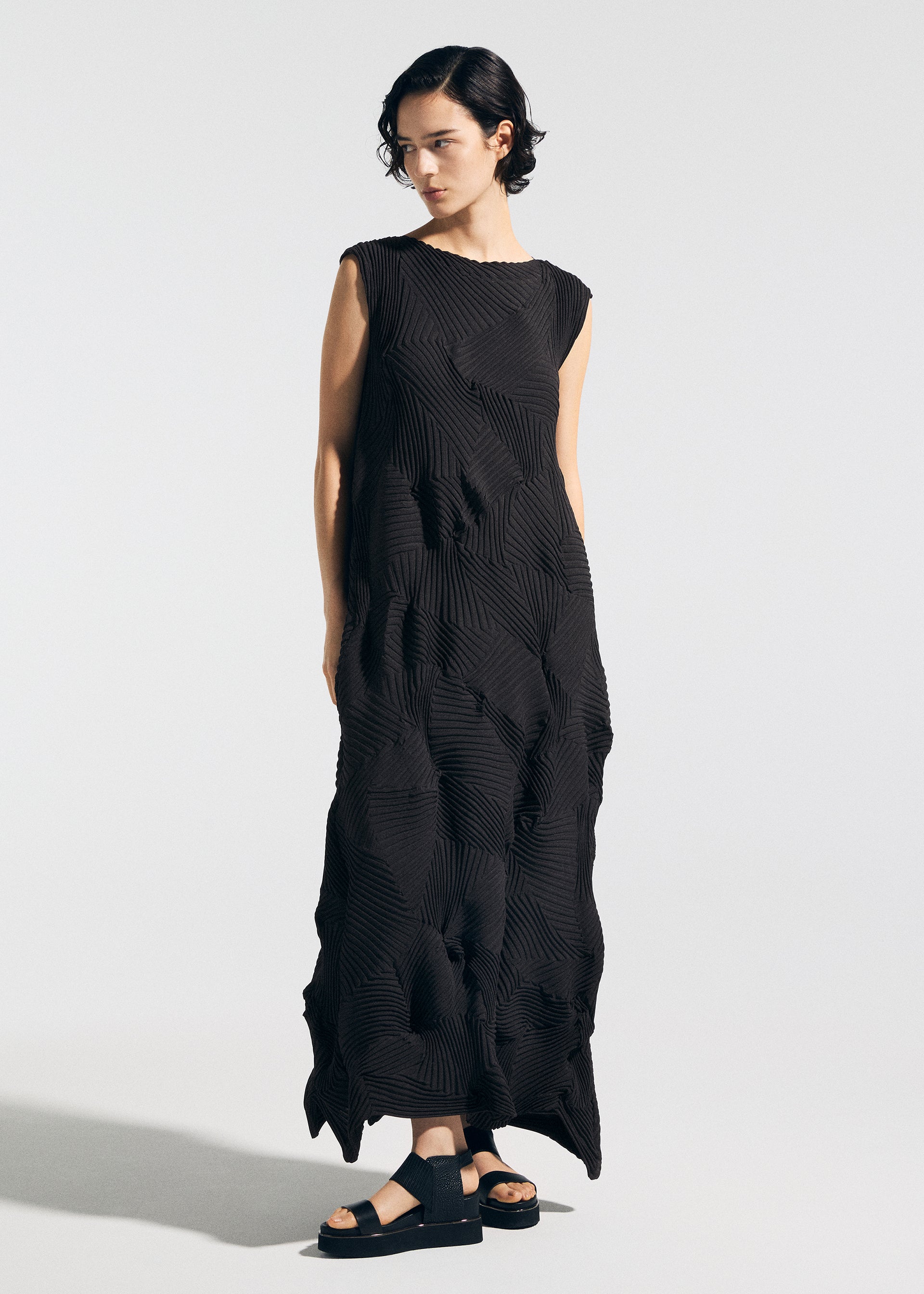 Zoomed view A person with short dark hair models the TYPE-P 003-1 dress by A-POC ABLE ISSEY MIYAKE— a sleeveless, pleated piece with textured geometric folds—paired with black sandals against a plain light background.