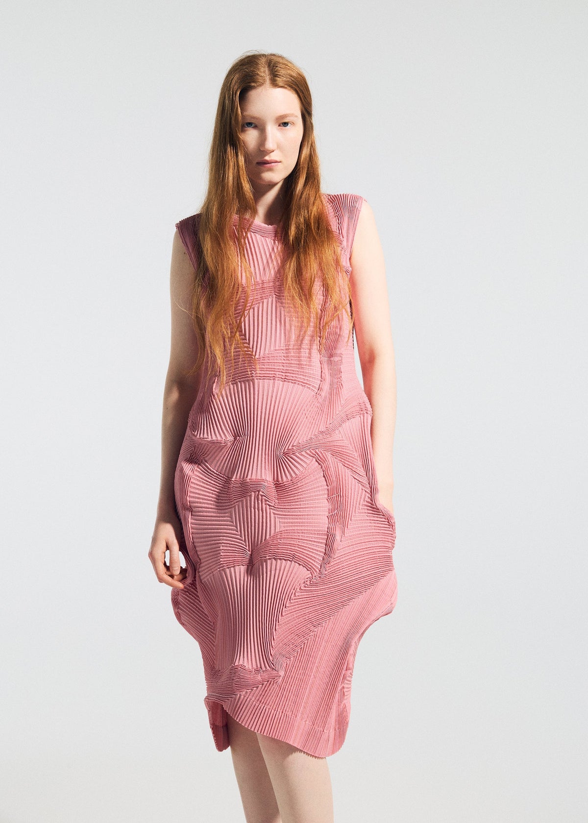 A woman with long red hair wears the TYPE-O 003 sleeveless, textured pink dress by A-POC ABLE ISSEY MIYAKE, featuring intricate geometric pleats. She stands against a plain light background, glancing slightly at the camera. view 6 of 7
