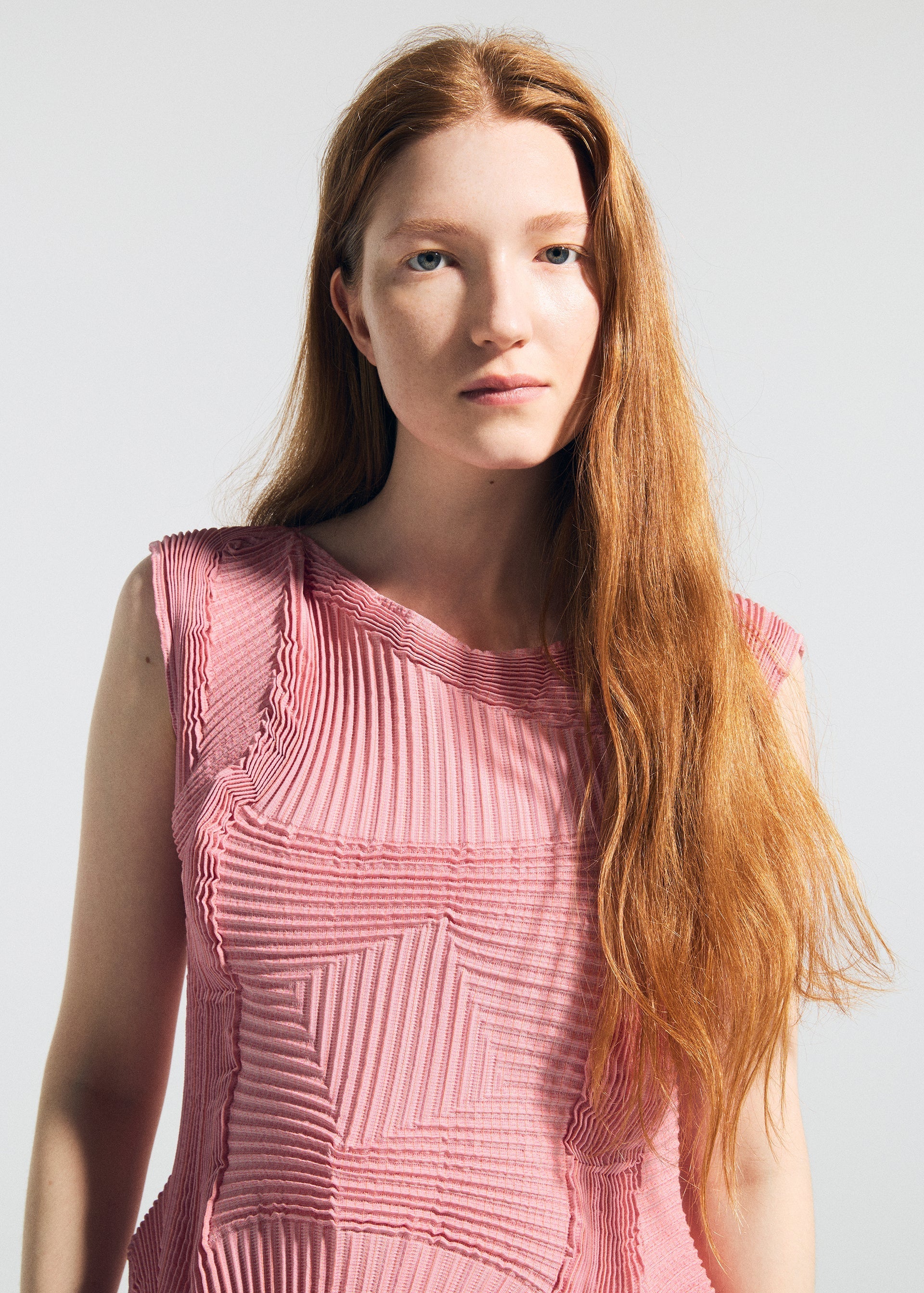 Zoomed view A woman with long red hair models the TYPE-O 003 BK sleeveless textured pink top by A-POC ABLE ISSEY MIYAKE, standing against a plain light background and looking calmly at the camera.