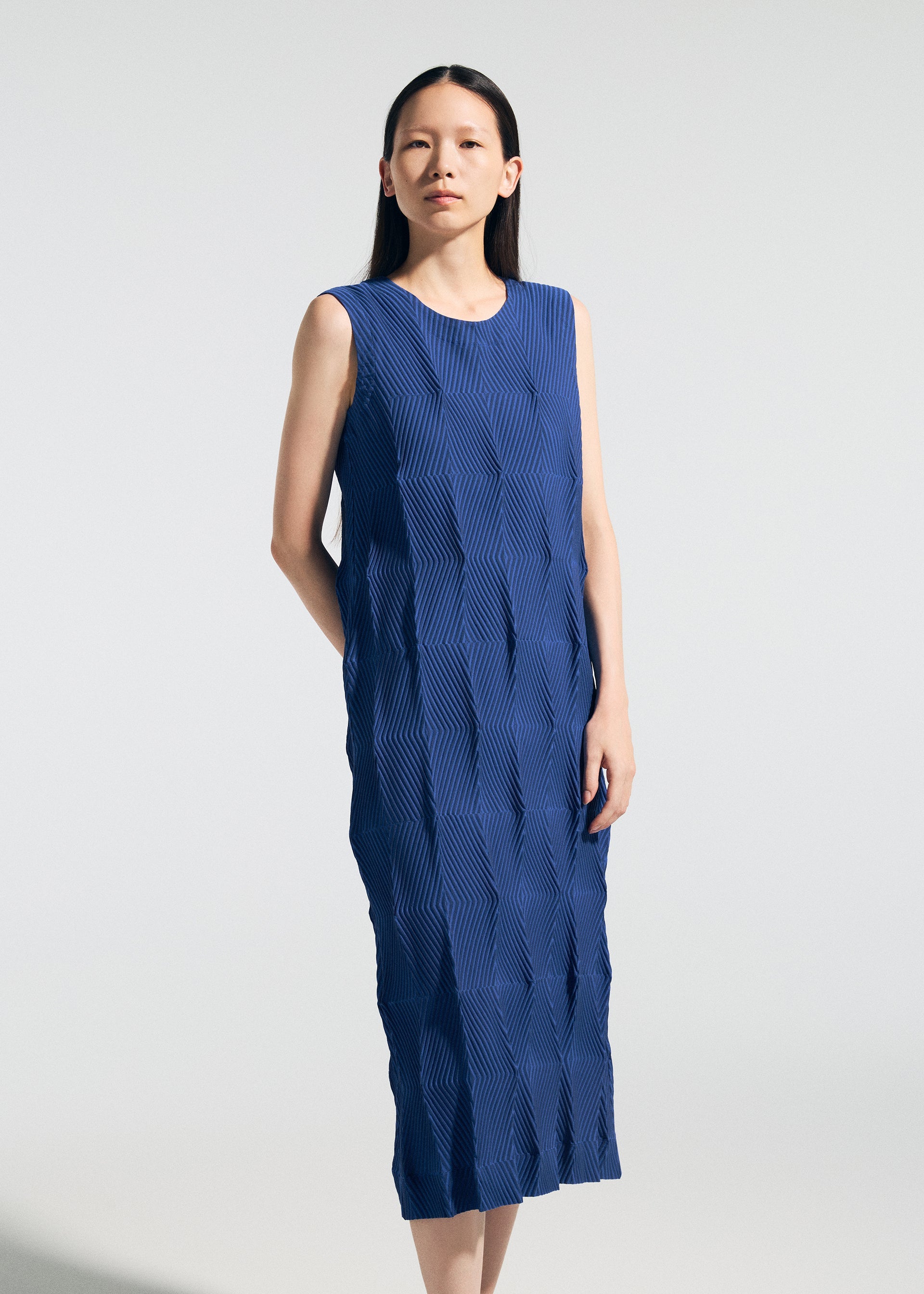 Zoomed view A woman stands against a plain light background, looking forward with her right hand behind her back, wearing the TYPE-W 009-2 dress from A-POC ABLE ISSEY MIYAKE—a sleeveless, ankle-length blue piece featuring a geometric pleated texture. Crafted from stretchy polyester for effortless movement and modern appeal, this dress offers a relaxed fit.