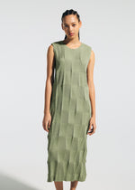 A person stands wearing the TYPE-W 009-2 dress by A-POC ABLE ISSEY MIYAKE—a sleeveless, knee-length green Steam Stretch dress featuring a geometric textured pattern. The relaxed-fit dress is set against a plain, light-colored background, and the person has a neutral expression.