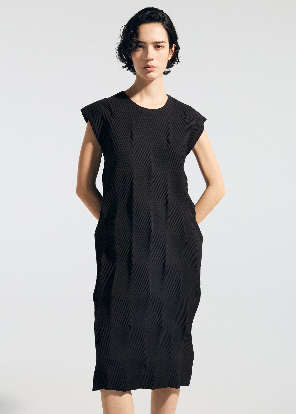 A person with short, dark hair stands against a plain background wearing the TYPE-W 009-1 sleeveless black dress by A-POC ABLE ISSEY MIYAKE. Their hands in the pockets highlight the relaxed fit, subtle geometric texture, and stretchy polyester comfort. view 5 of 5